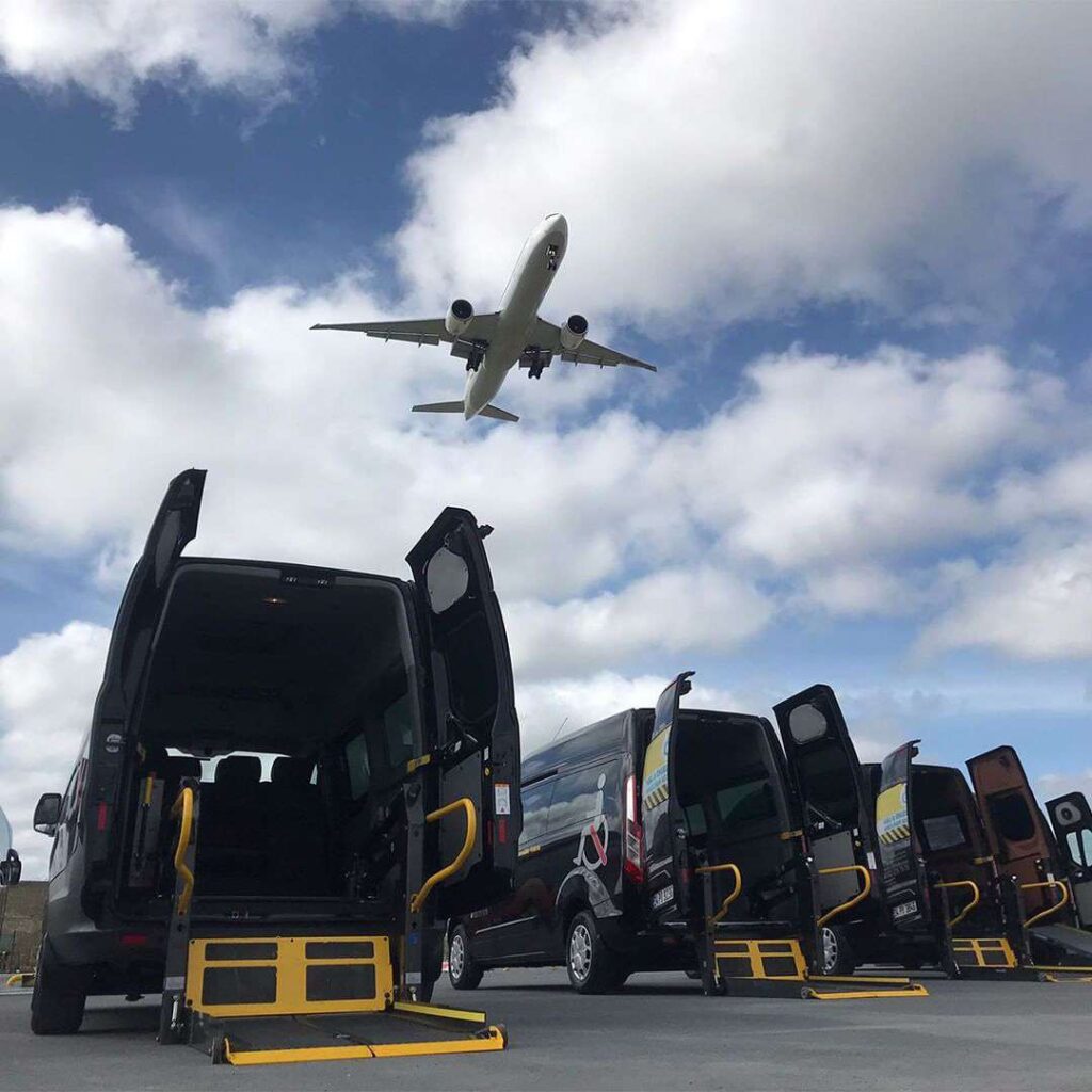 Row of ambulette service vans with wheelchair lifts extended, parked under a plane approaching for landing, highlighting airport accessibility